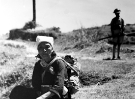 A woman in Yunnan province, China, rests to eat a meal, during the hard days of WWII.  Note the foot container on her lap and the chopsticks in her hands. A soldier with gun stands in the background.  From the collection of Eugene T. Wozniak.