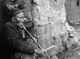 Local people in Yunnan province, China: Elderly man sits in village doorway and smokes from very long pipe. During WWII.