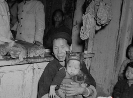 A woman holds a small child in a shop in Yunnan, China, during the Second World War.  Note the straw sandals hanging on the shop window in the back.